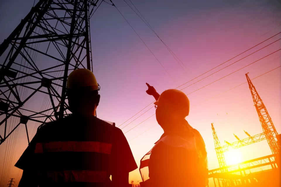 Two utility workers looking up at power lines against purplish sunset sky.